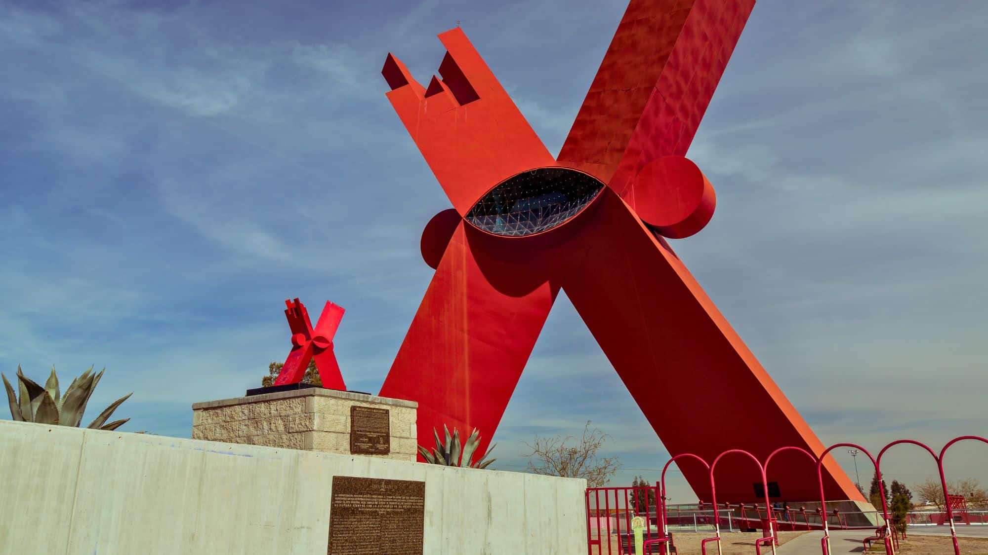 The large red "X" monument in Ciudad Juárez stands tall against a clear blue sky. The modern, geometric structure dominates the scene, symbolizing the city's identity and culture.
