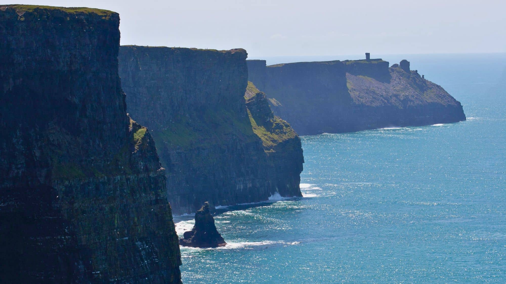 The rugged Cliffs of Moher tower over the deep blue ocean, their sheer faces covered in patches of green. The horizon stretches into the distance under a clear sky.