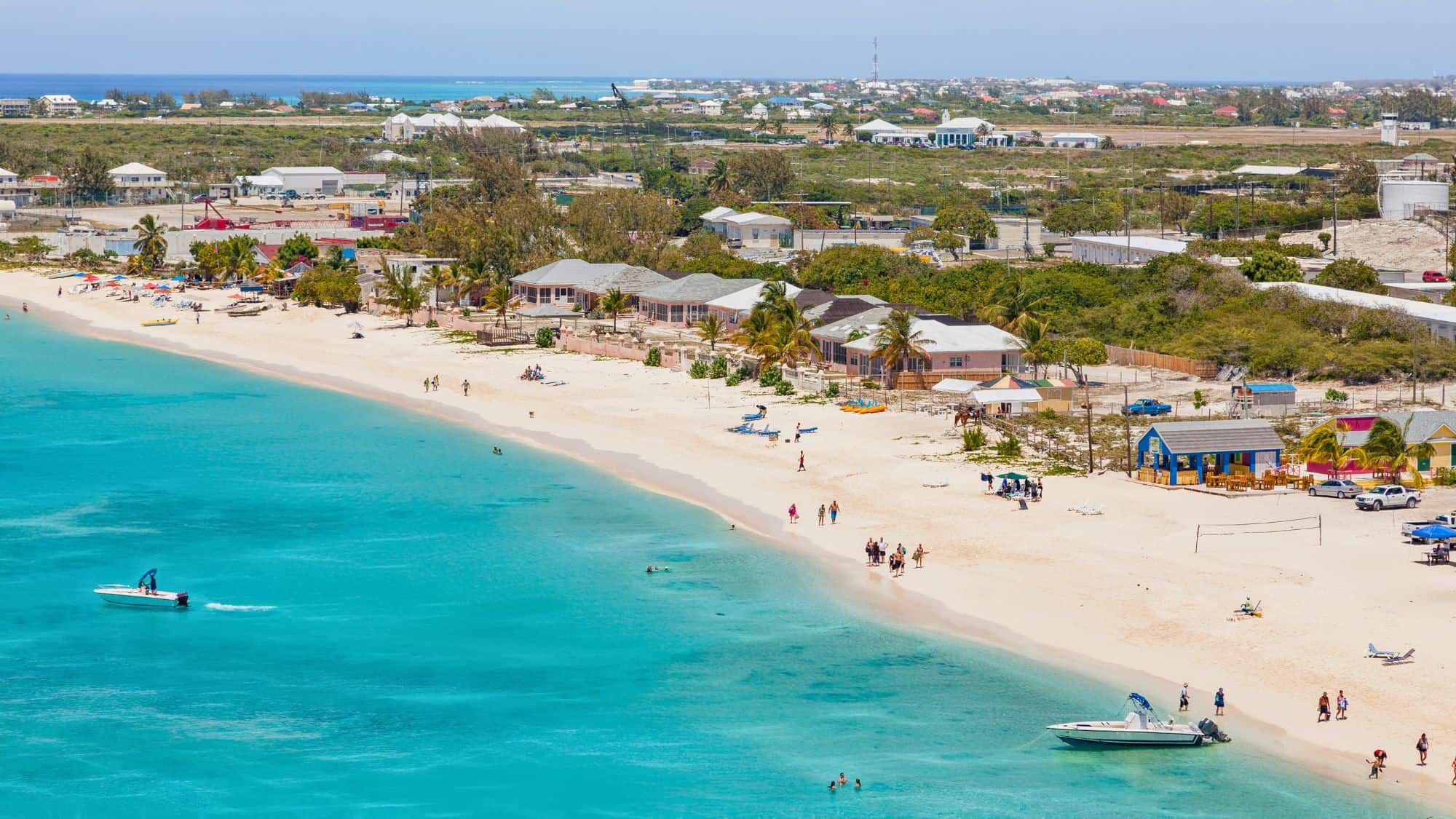 A pristine beach with turquoise waters gently meeting the white sandy shore, lined with colorful buildings and beach umbrellas. Small boats float in the calm sea, while visitors stroll along the beach and relax near the water.