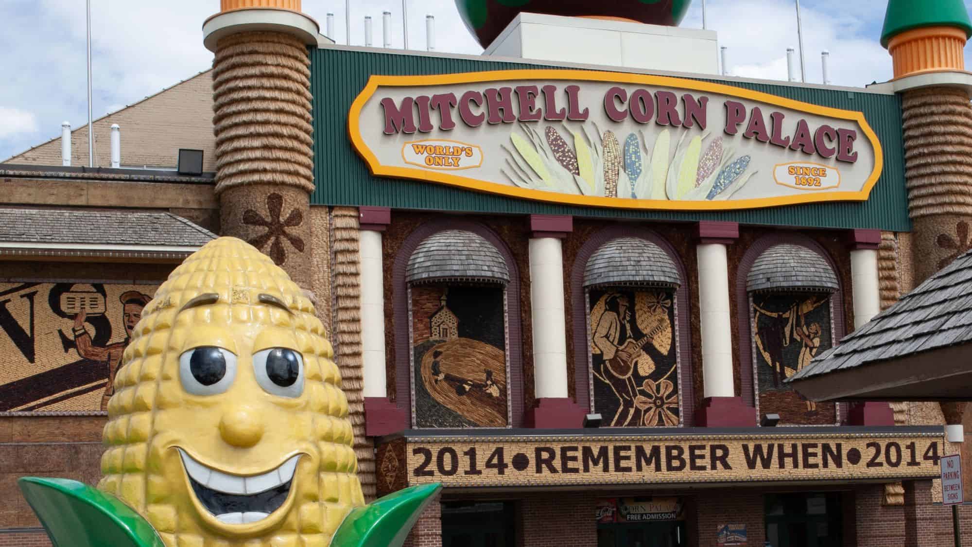 The front of the Mitchell Corn Palace, adorned with intricate corn murals and a large sign reading “MITCHELL CORN PALACE World's Only Since 1892.” A giant, cartoonish ear of corn statue with a smiling face stands in front, welcoming visitors.