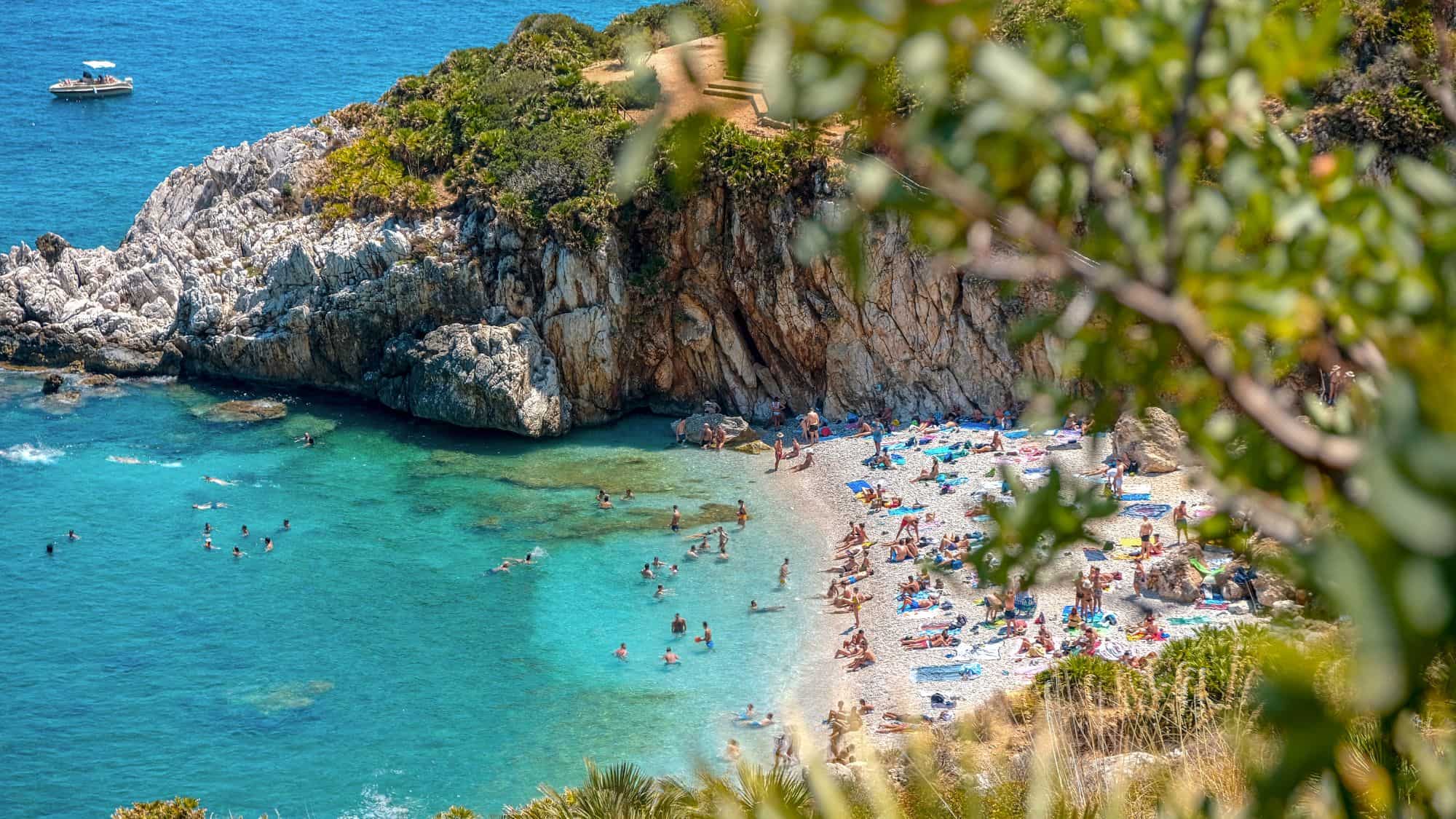 A hidden cove within the Zingaro Nature Reserve, Sicily, featuring crystal-clear water, rugged cliffs, and sunbathers along the rocky beach. Swimmers enjoy the shallow turquoise waters near the shore.