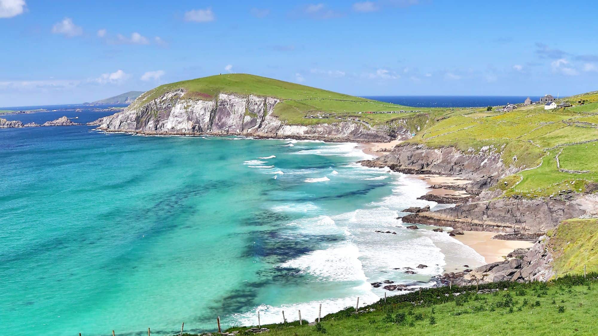 A stunning view of the Dingle Peninsula in Ireland, showcasing turquoise waters crashing against rugged cliffs, with rolling green hills stretching into the distance under a bright blue sky.