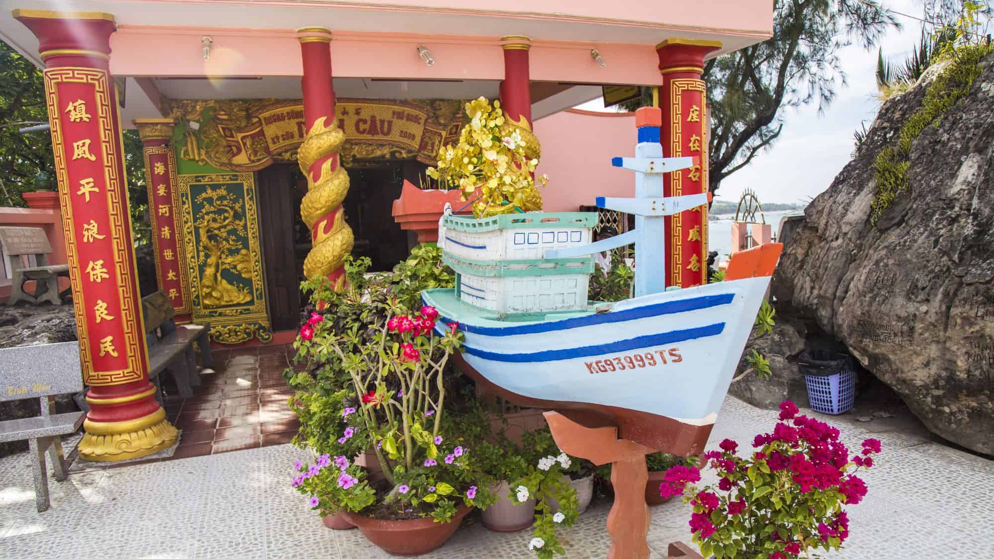 A vibrant Vietnamese temple featuring red and gold pillars, floral decorations, and a small blue and white model boat at the entrance, symbolizing maritime protection.