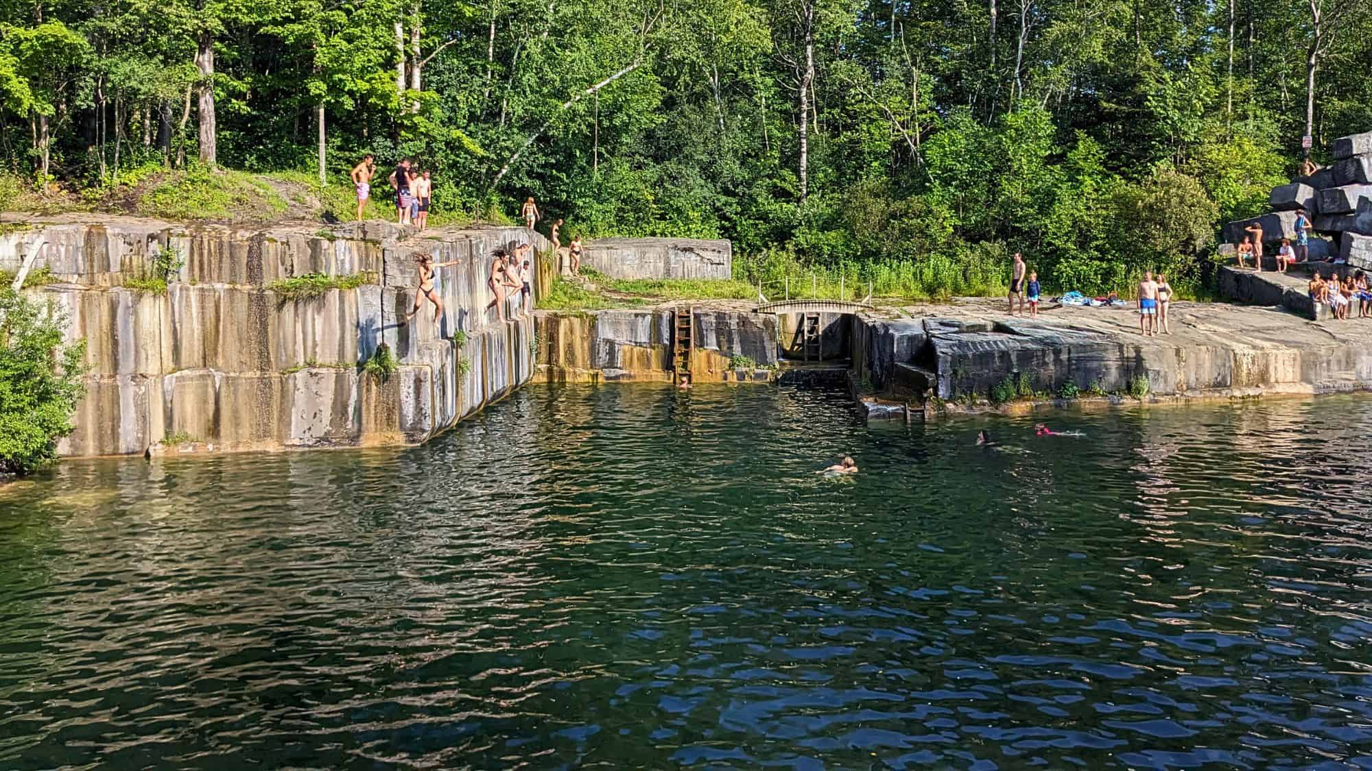 People gather at the Dorset Marble Quarry, some perched on stone ledges while others leap into the deep, clear water below. The rugged, squared-off marble walls contrast with the surrounding greenery.