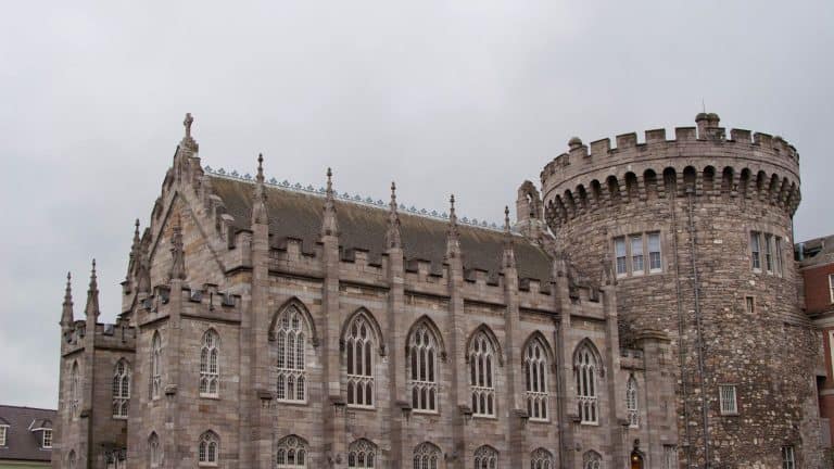 A detailed view of Dublin Castle in Ireland, featuring the Gothic Revival Chapel Royal alongside the medieval Record Tower. The gray stone facade, intricate windows, and battlements stand under an overcast sky.