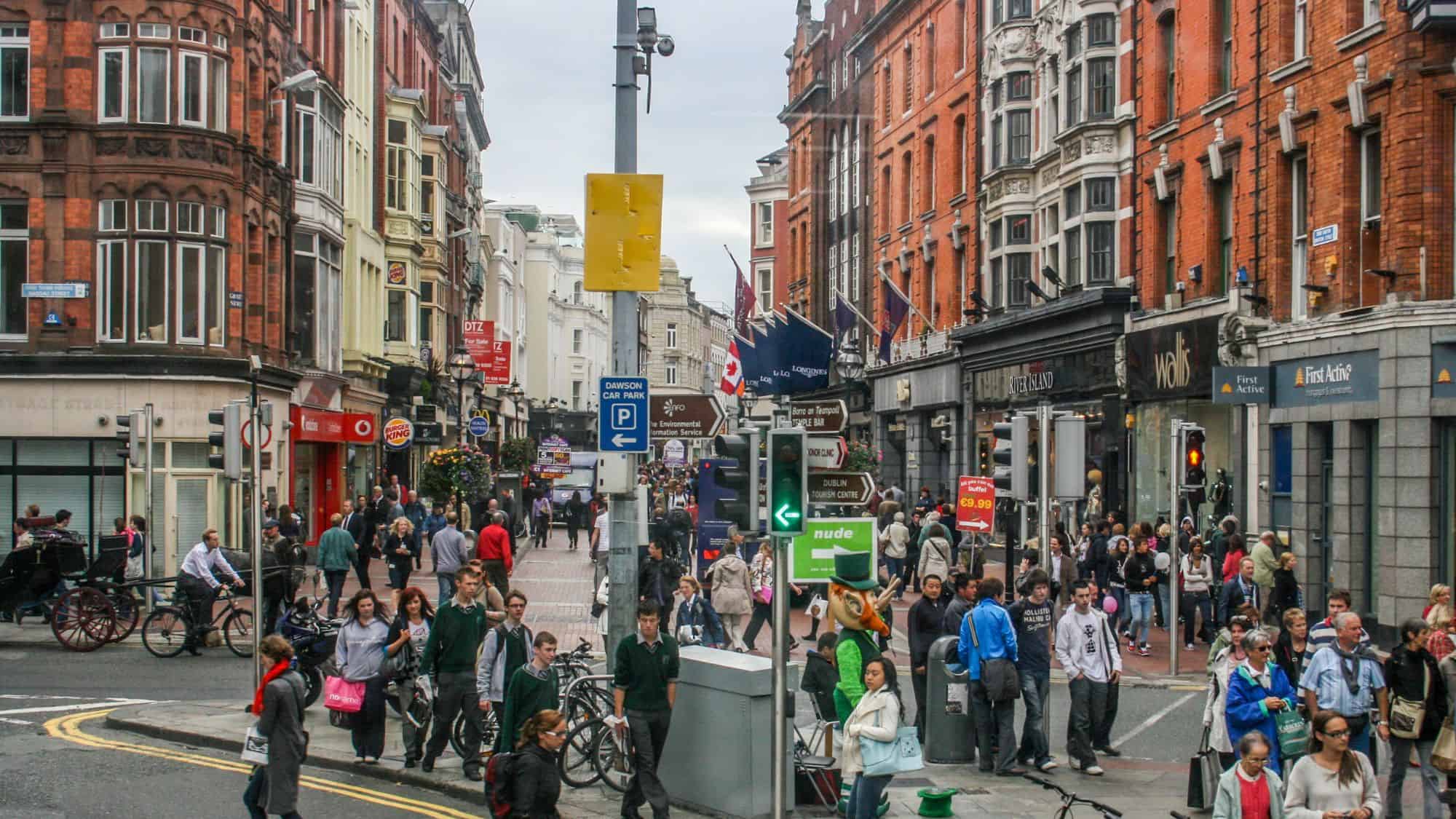 A busy street scene in Dublin, Ireland, with pedestrians walking along historic red-brick buildings housing shops, restaurants, and pubs. The street is alive with shoppers, tourists, and street performers under a cloudy sky.