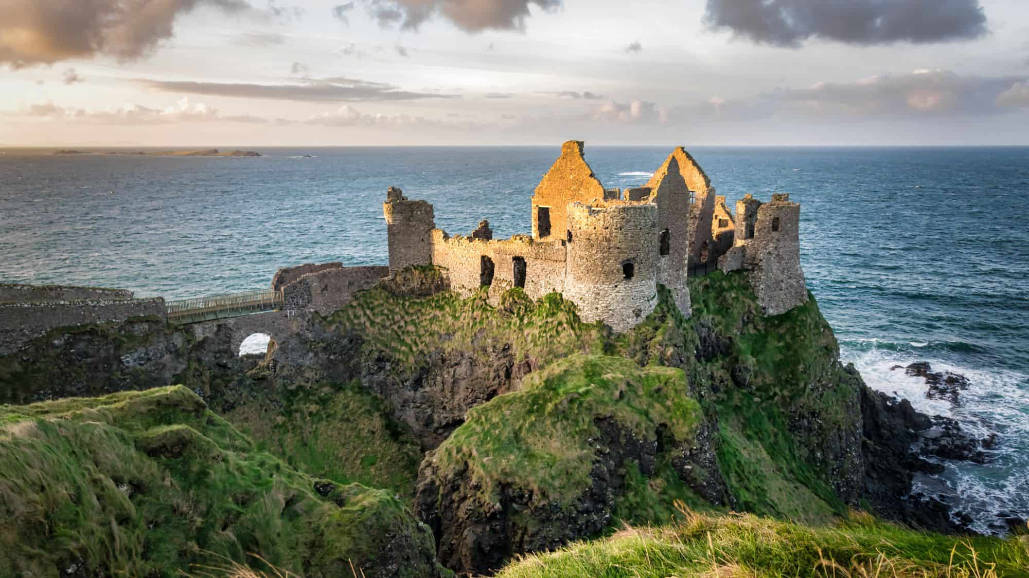 The ruins of Dunluce Castle sit dramatically on a cliffside overlooking the ocean. The stone walls, some crumbled, contrast with the vibrant green grass and the sea under a partly cloudy sky.