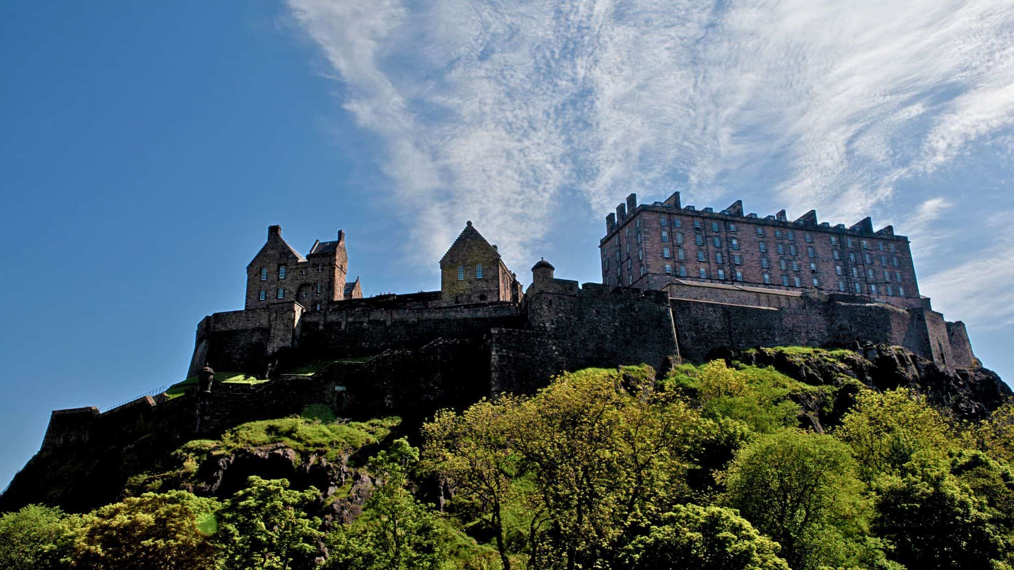 Edinburgh Castle sits atop Castle Rock, its stone walls blending with the rugged cliffside. The fortress is surrounded by green trees under a bright blue sky with wispy clouds.