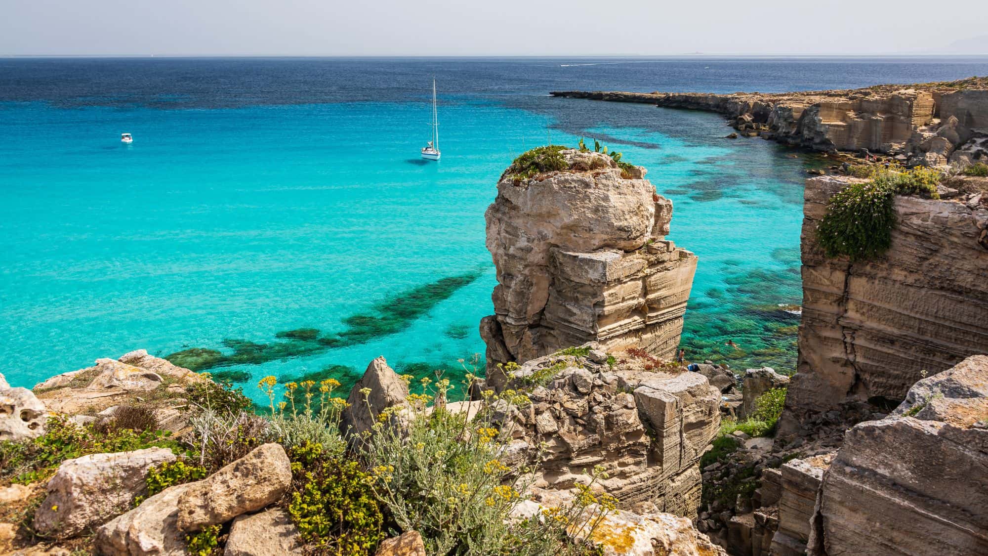 A breathtaking view of the turquoise waters surrounding the rocky cliffs of Favignana, Sicily. A sailboat floats in the distance, adding to the tranquil, sunlit seascape.