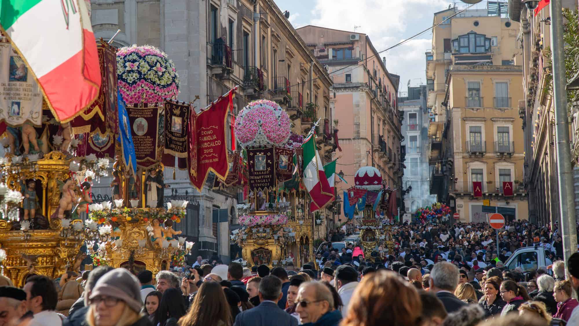 A lively religious festival in Catania, Sicily, with large crowds gathered in the streets. The event features ornate religious floats adorned with flowers, banners, and statues, while participants wave Italian flags and celebrate in traditional attire.