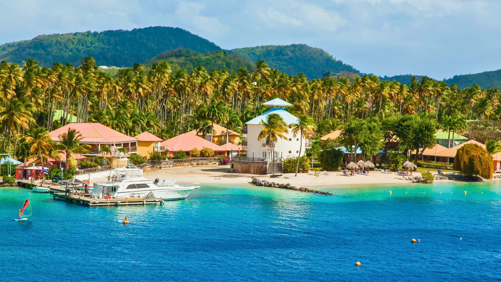 A picturesque Caribbean beach scene with colorful buildings, palm trees, and yachts docked along the shore, set against green hills in Fort-de-France, Martinique.