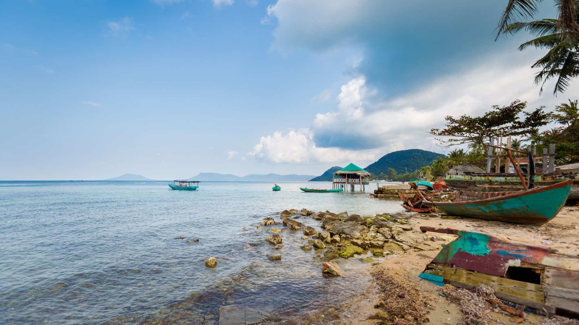 A serene beach scene with clear blue waters, rustic fishing boats, and weathered wooden structures along the shore, with distant islands visible on the horizon.