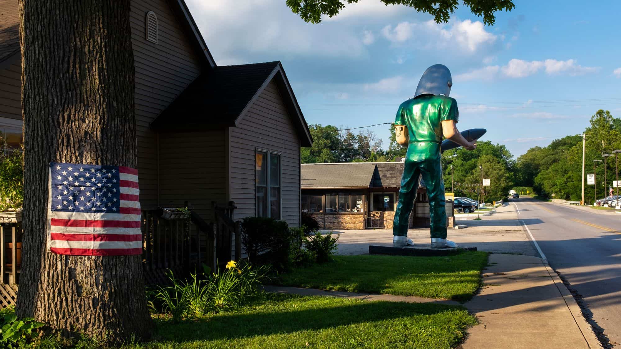 A towering statue of a green space-age figure holding a silver rocket, standing on a sidewalk along an old highway. The figure, known as the Gemini Giant, is a famous roadside Muffler Man attraction in Illinois.