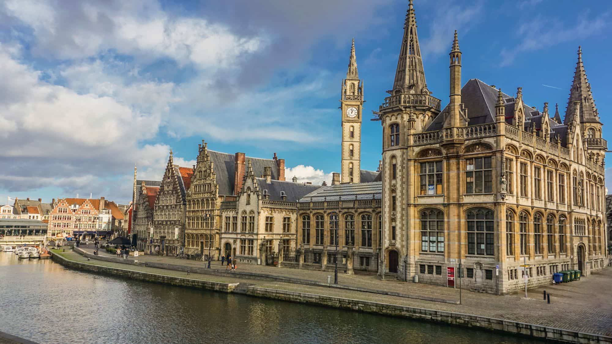 A scenic canal lined with historic Flemish buildings featuring ornate gables and a clock tower. Reflections of the architecture shimmer in the water, adding to the old-world European ambiance.
