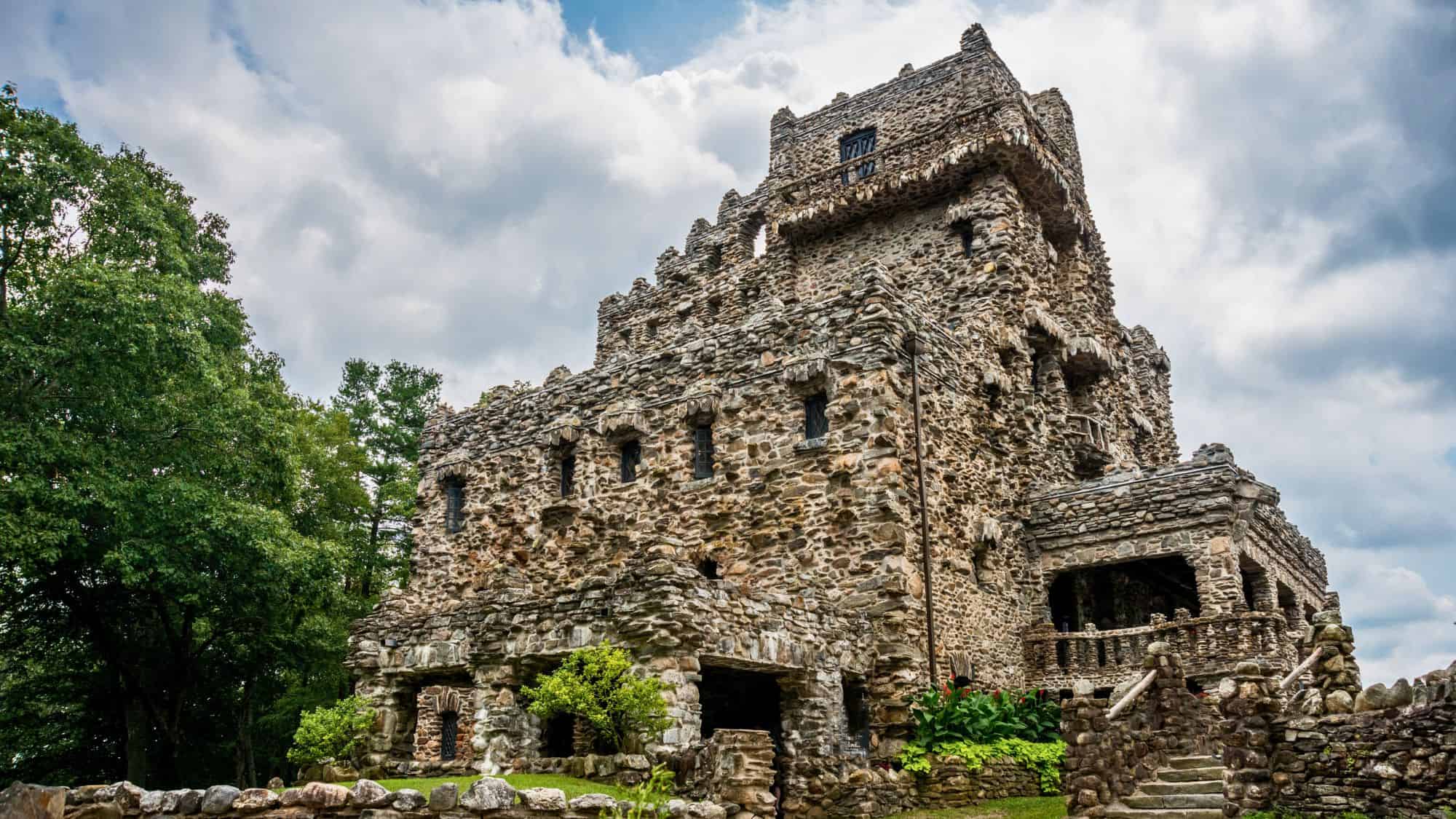 A rugged stone castle with an unusual, whimsical design stands surrounded by lush greenery. The structure's intricate stonework and turrets give it a fairytale-like appearance, set against a cloudy sky.