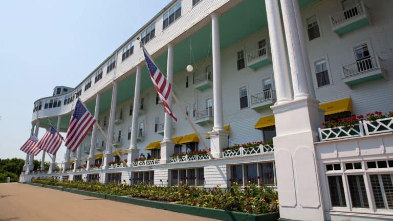 A historic white hotel with towering columns and American flags lining the front, featuring yellow awnings and vibrant flower beds along the entrance. The classic architecture and long porch showcase the elegance of this landmark destination.