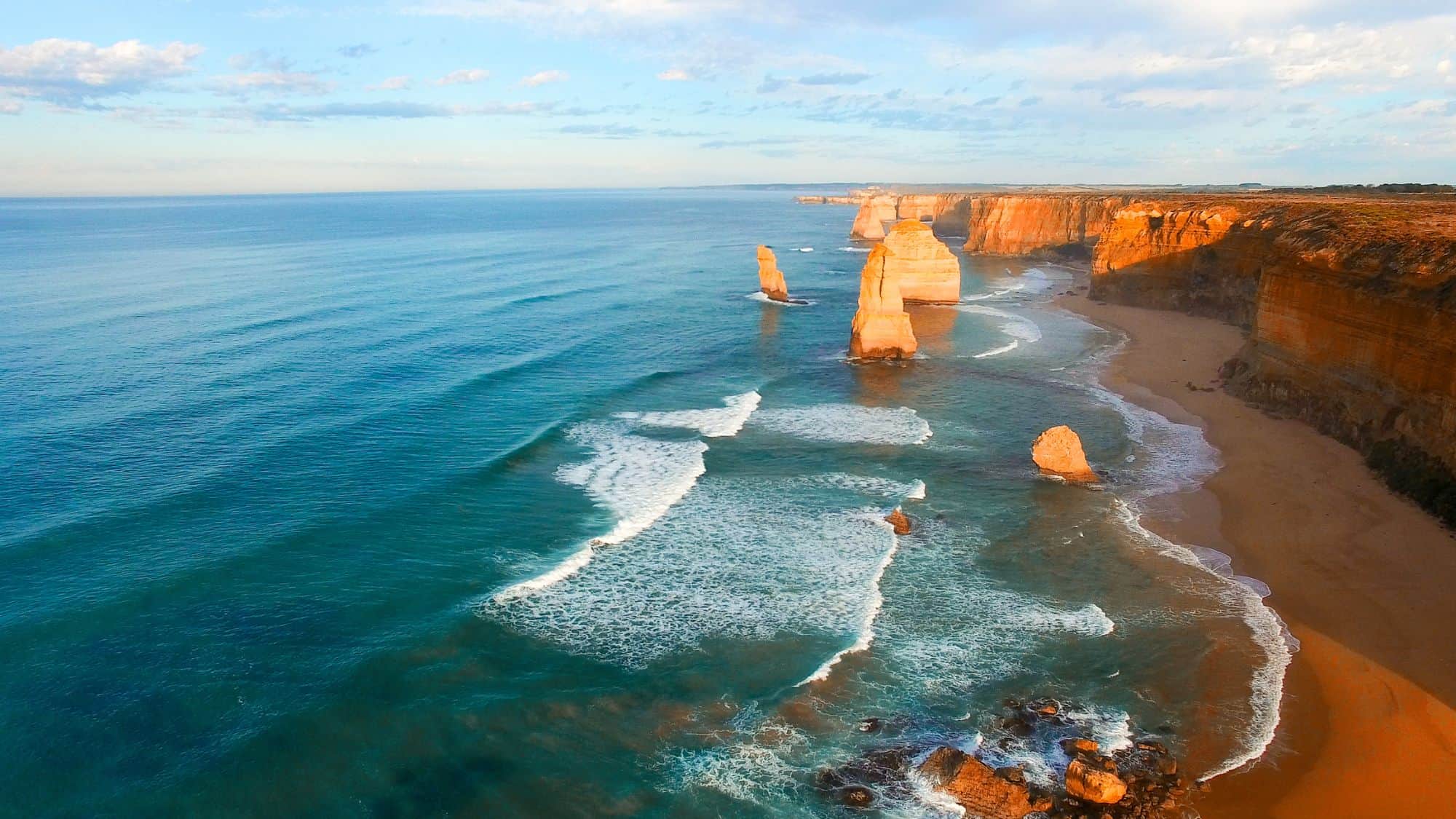 A panoramic view of the Twelve Apostles along Australia's Great Ocean Road, with limestone stacks rising from the turquoise waters, golden sand beaches, and dramatic cliffs illuminated by the soft light of sunset.