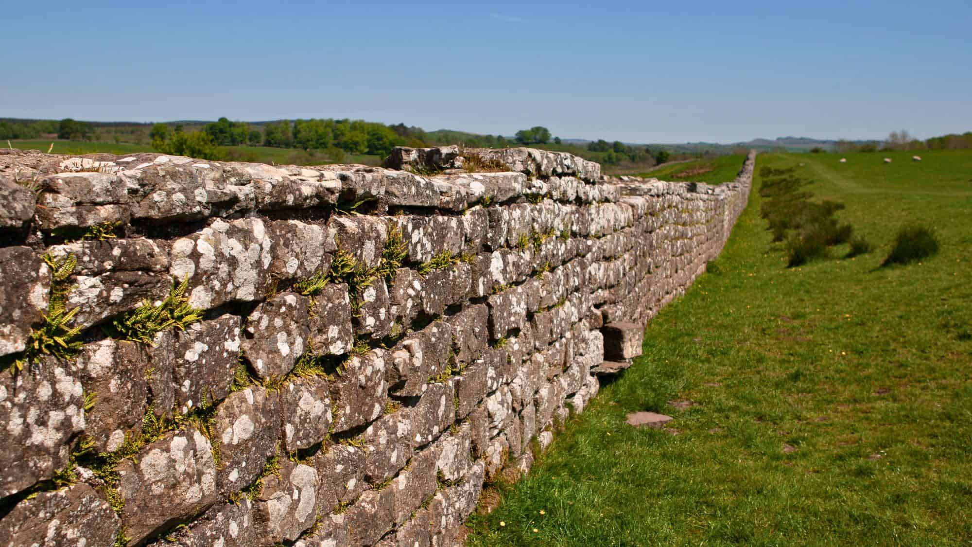 An ancient stone wall stretches across a grassy landscape, weathered and covered with moss. The wall extends into the horizon under a bright blue sky.