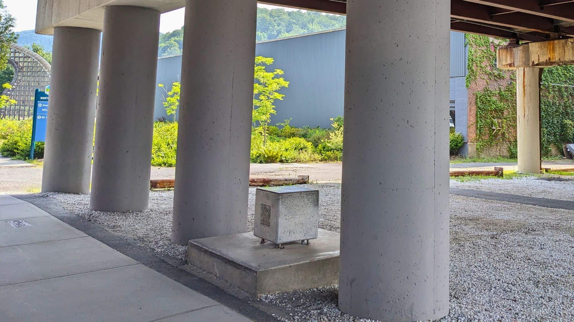 Beneath a concrete overpass, a small metal cube rests on a pedestal, known as the Harmonic Bridge. The industrial setting contrasts with the natural greenery in the background.