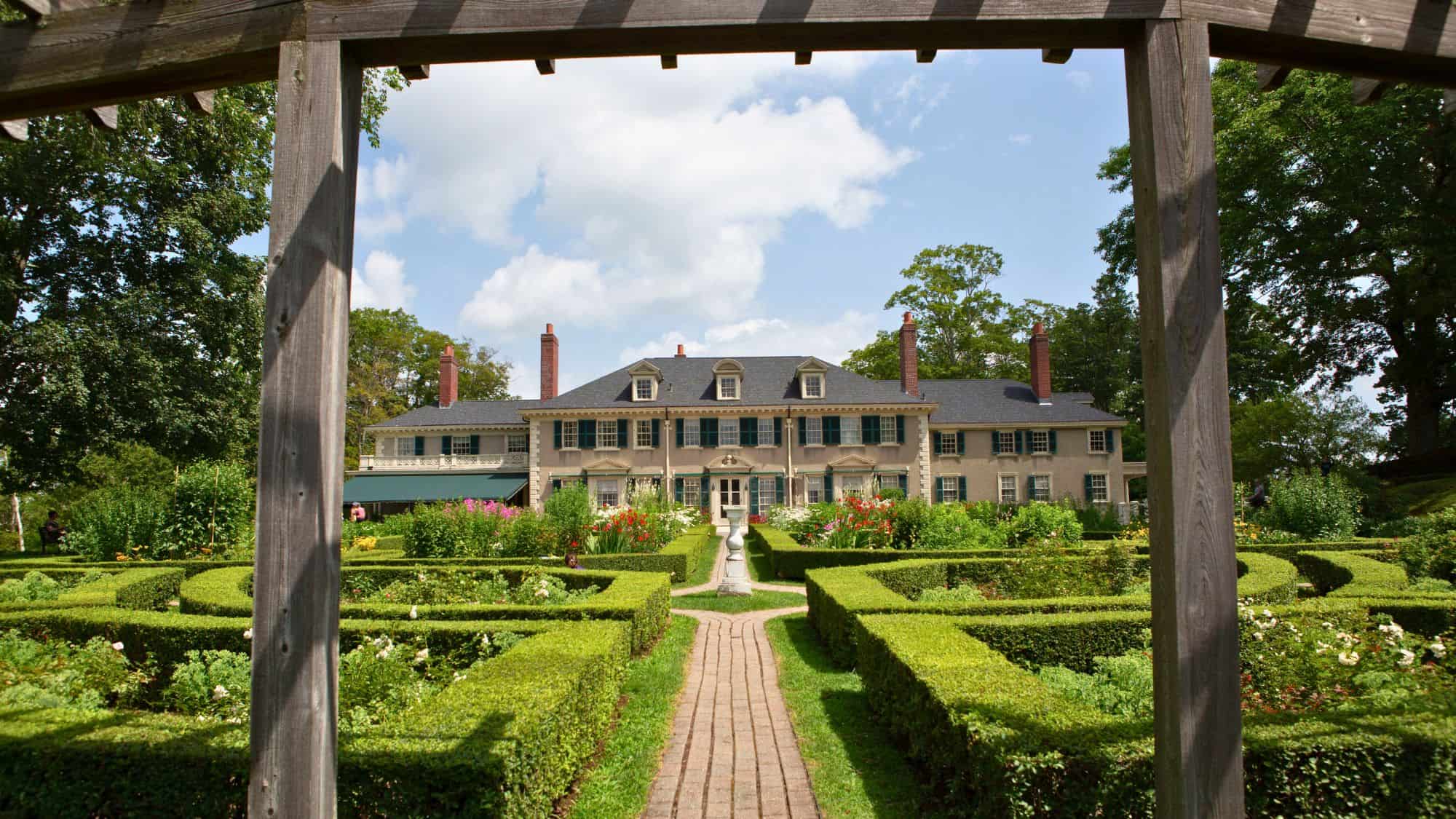 A stately mansion with a symmetrical design sits beyond perfectly manicured gardens at Hildene. Boxwood hedges form intricate patterns leading up to the grand entrance, framed by a wooden pergola.