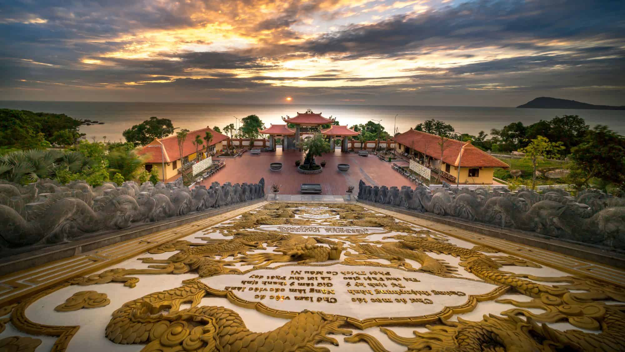 A stunning Buddhist temple overlooking the ocean, with an intricately designed golden dragon staircase leading to a courtyard at sunset.