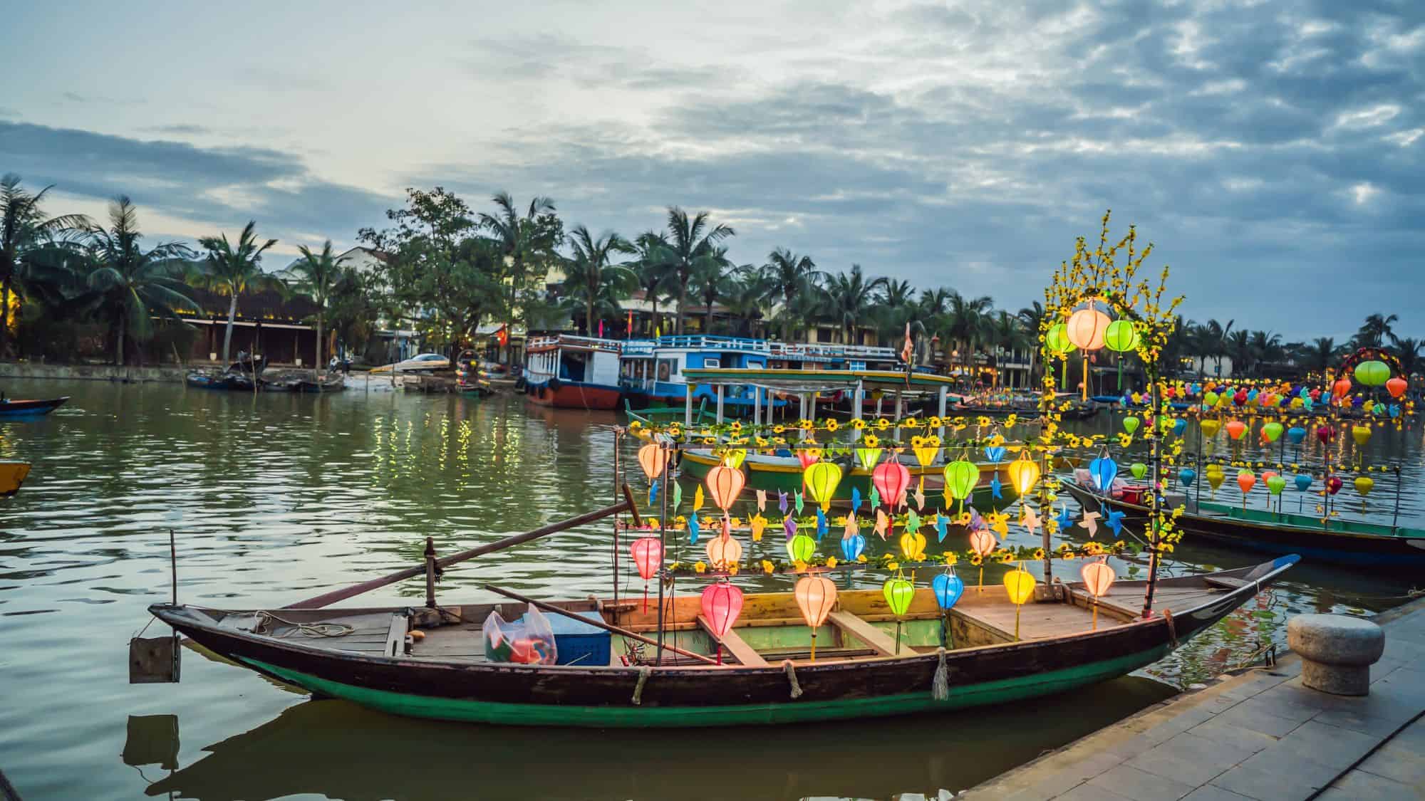 A riverboat decorated with colorful lanterns floats on calm waters at dusk, with more illuminated boats and palm trees in the background. The warm glow of lanterns reflects on the water, creating a magical atmosphere in this ancient town.