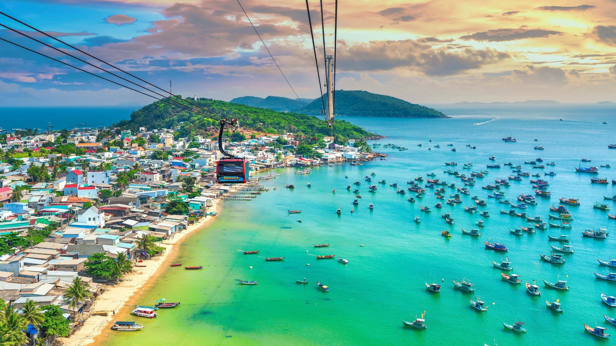 A scenic aerial view of a coastal fishing village with colorful houses, turquoise waters filled with boats, and a cable car gliding above, set against lush green hills.