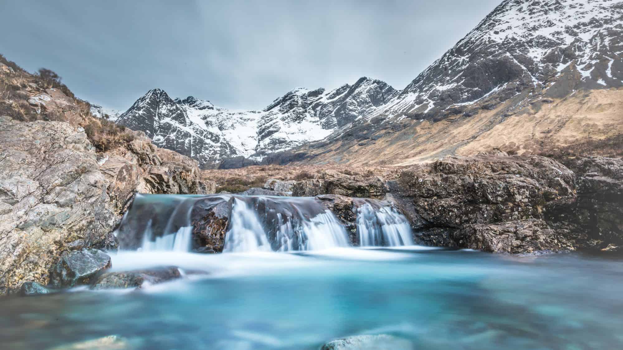 A turquoise waterfall cascades into a crystal-clear pool, surrounded by rocky terrain. Snow-capped mountains rise in the background under a soft, cloudy sky.