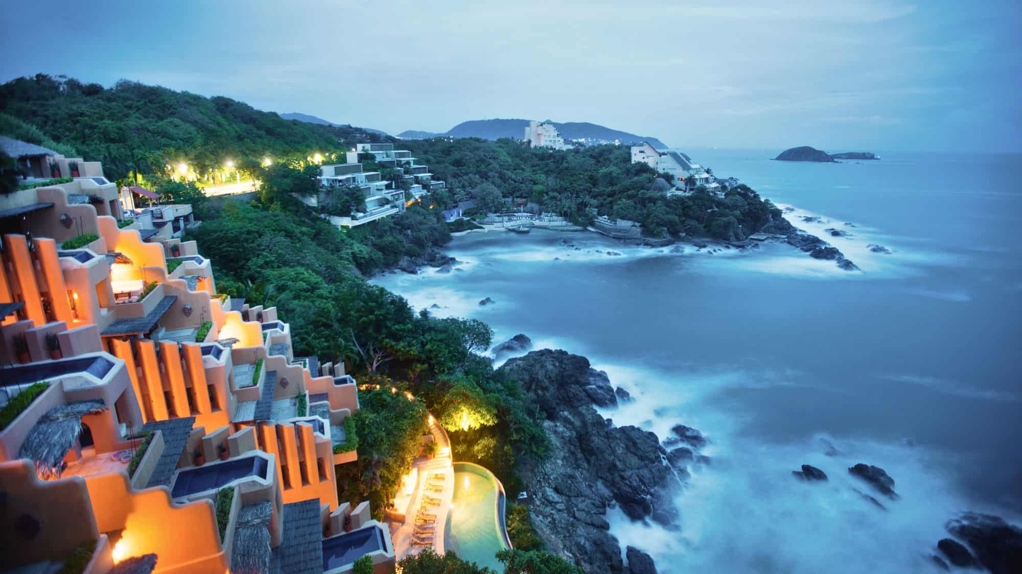A stunning dusk view of Ixtapa-Zihuatanejo, with luxurious terraced resorts glowing in warm light against a lush green hillside. Below, the ocean waves crash onto the rocky coastline, creating a dreamy, misty effect.