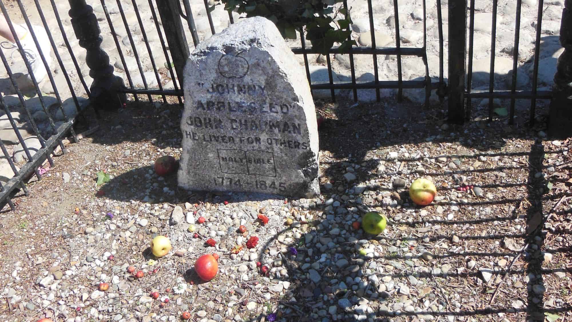 A simple, weathered gravestone marking the final resting place of Johnny Appleseed (John Chapman), surrounded by a black metal fence. Apples and flowers are scattered at the base as tributes from visitors.