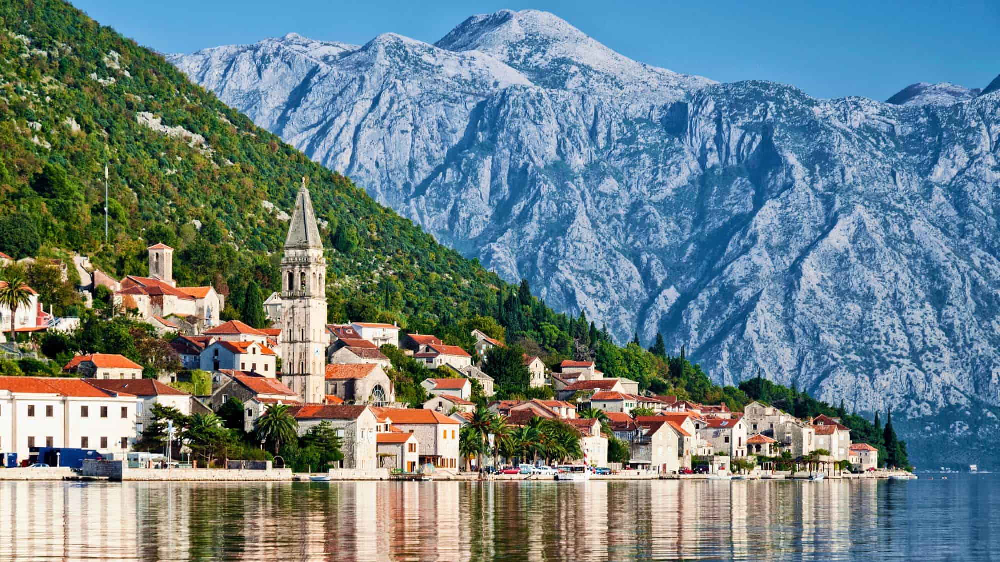 A charming coastal town nestled between towering mountains and the shimmering waters of the Bay of Kotor. Red-roofed stone buildings and a church steeple stand out against the dramatic rocky landscape.