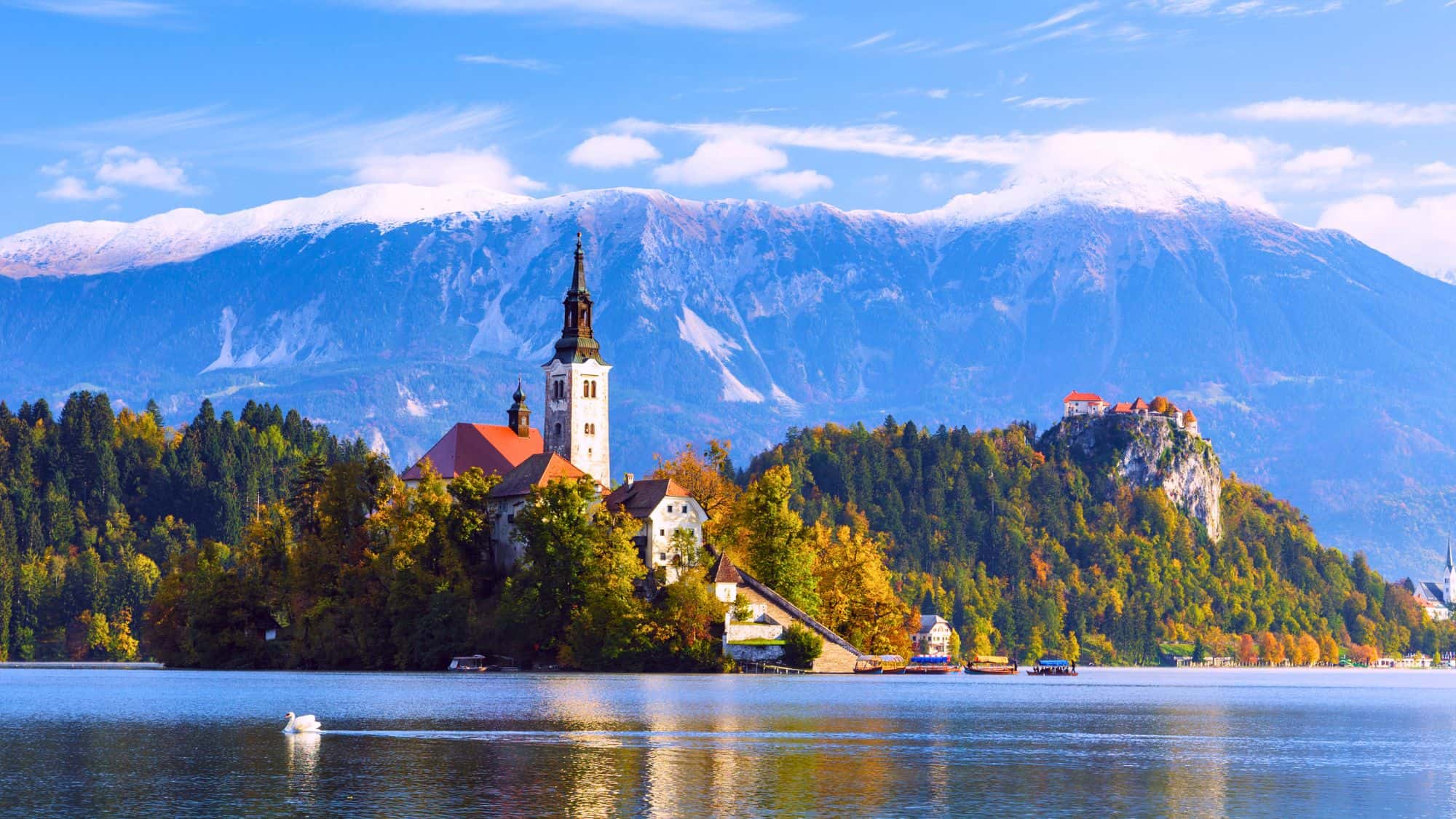 A fairytale-like church sits on a small island in the middle of a tranquil lake, surrounded by autumn foliage. Snow-capped mountains rise in the background, while the still waters reflect the stunning scenery.