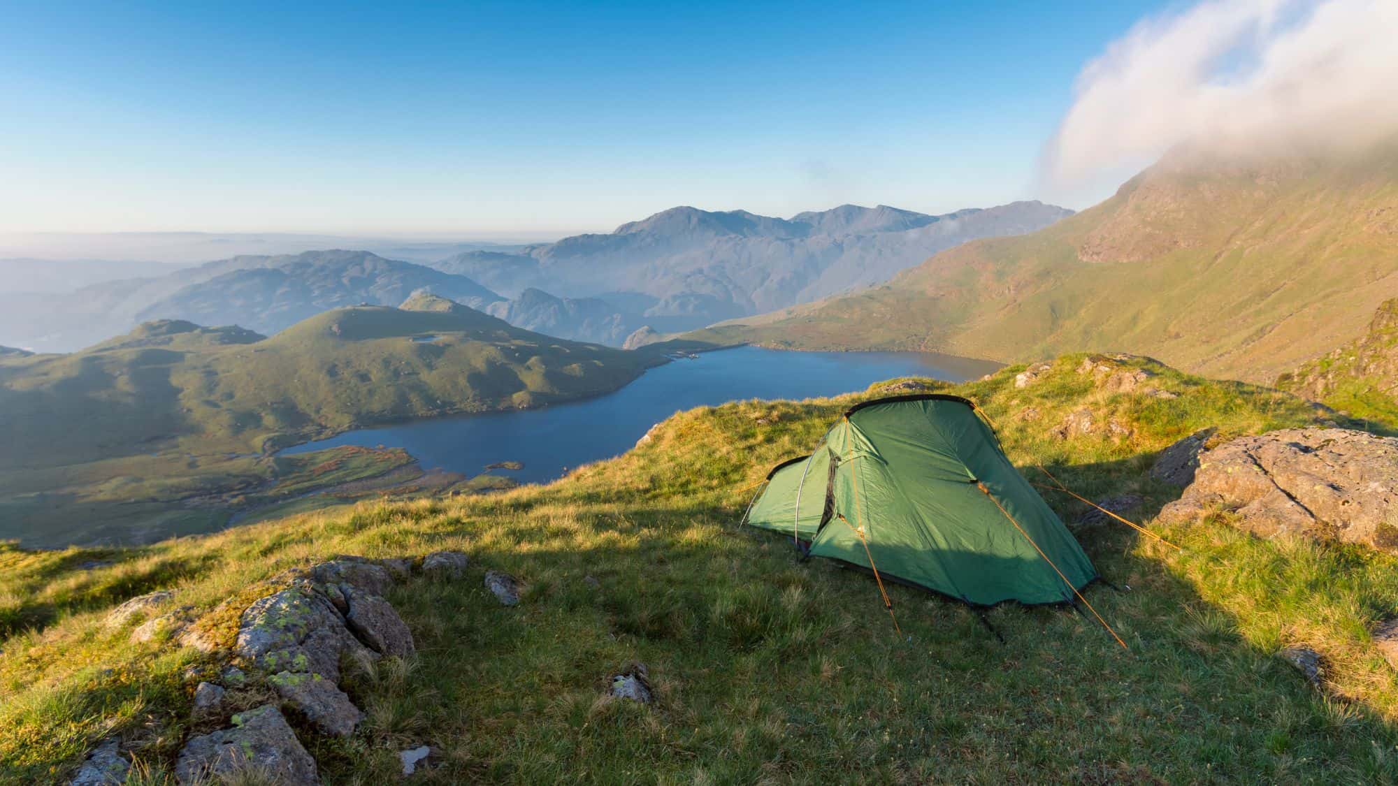 A peaceful camping scene with a green tent pitched on a grassy hill overlooking a lake and rolling hills, bathed in soft morning light with a low cloud brushing the mountain peaks in the distance.