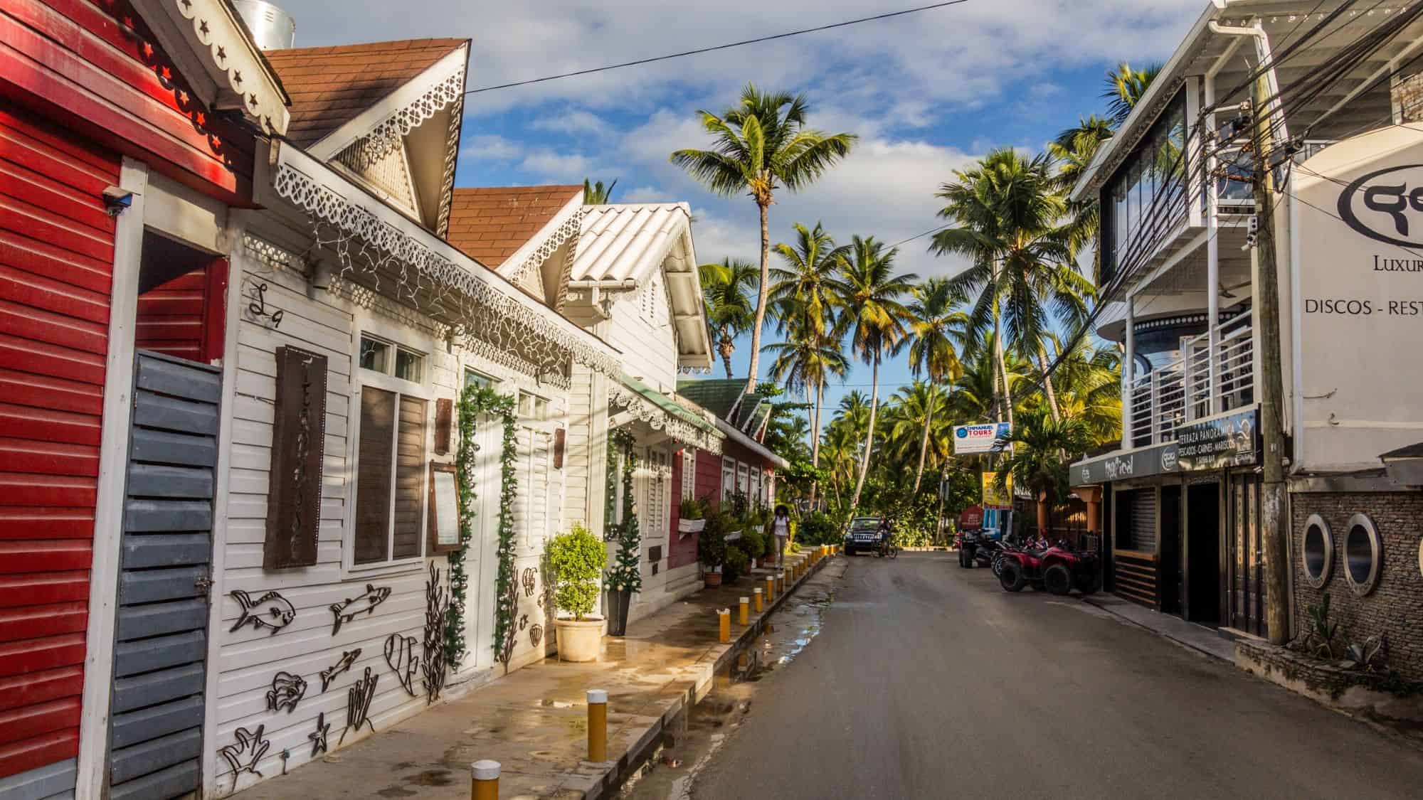 A charming Caribbean street lined with colorful wooden houses and palm trees, with a mix of small shops and restaurants. The relaxed tropical vibe is accentuated by the bright colors, local signage, and the clear blue sky.