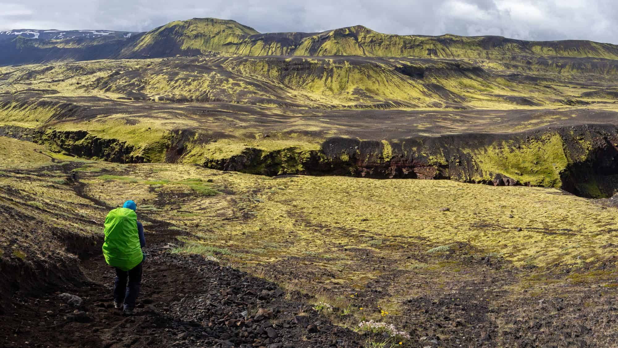 A vast Icelandic landscape featuring volcanic hills covered in vibrant green moss, dark lava fields, and a lone hiker with a bright green backpack walking along a rugged trail under a cloudy sky.