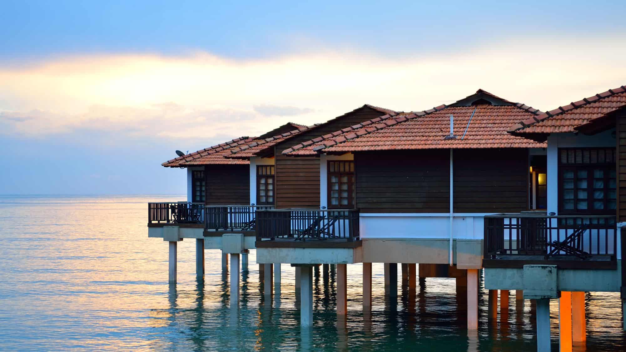 A row of wooden overwater bungalows with terracotta-tiled roofs stands against the backdrop of a calm ocean. The soft glow of the setting sun reflects off the water, adding to the serene atmosphere.