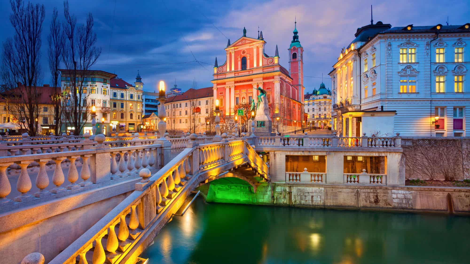 A twilight view of Ljubljana’s iconic pink Franciscan Church and triple bridge, illuminated against the deep blue sky. The city’s romantic riverfront glows with warm streetlights and reflections in the water.