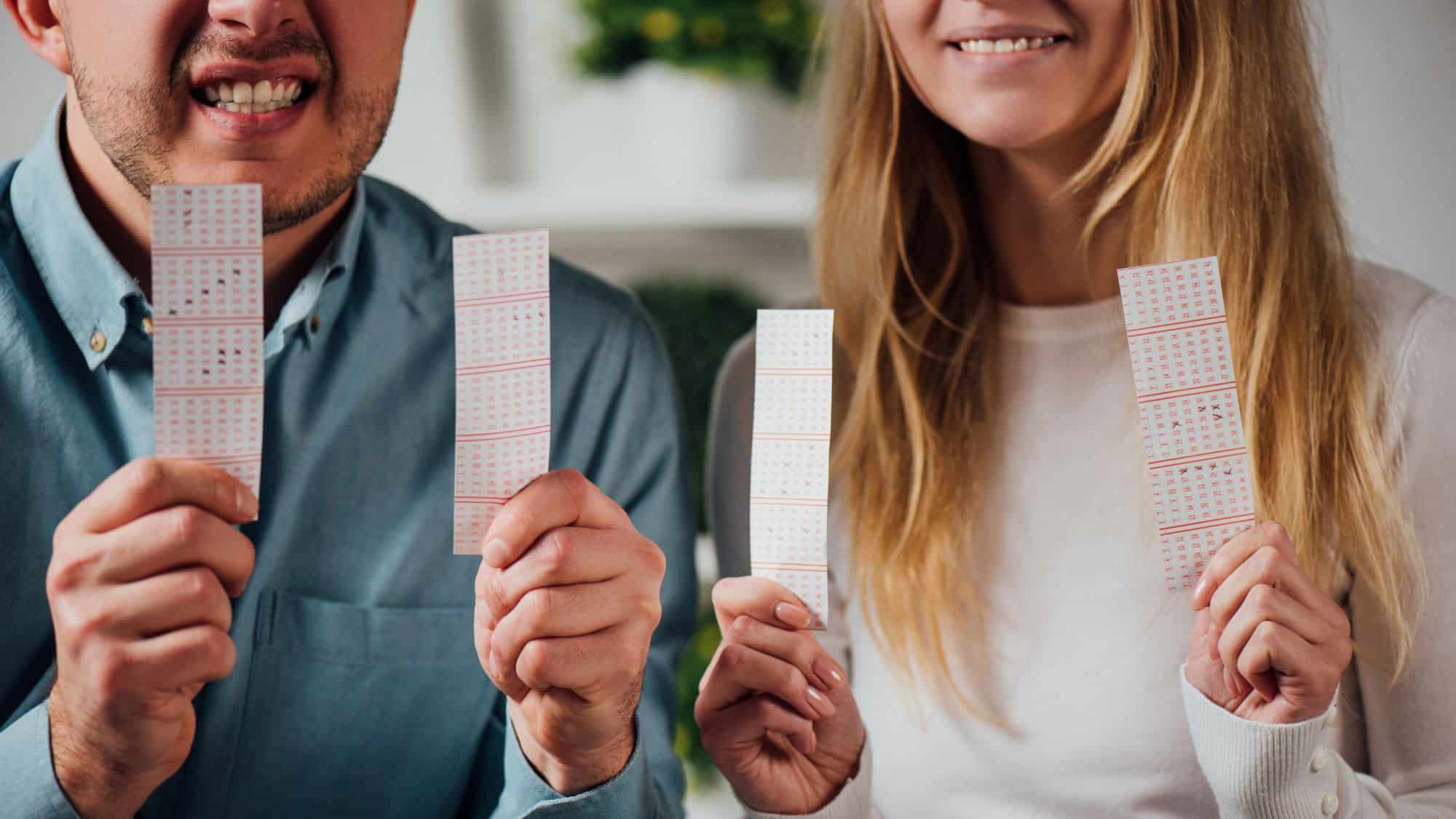 A man and woman holding several lottery tickets with excited and hopeful expressions. The tickets are marked with numbers, indicating they’ve played multiple rounds.