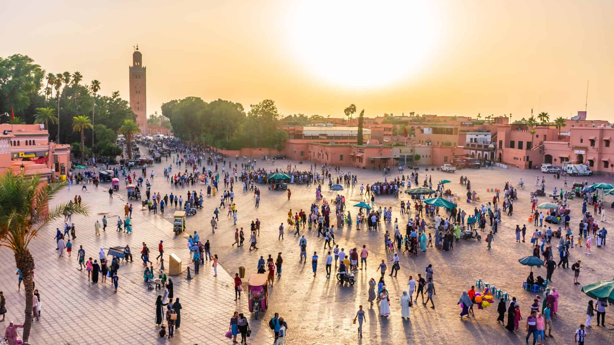 An aerial view of the famous Jemaa el-Fnaa square in Marrakech, Morocco, bustling with market stalls, performers, and locals. The towering Koutoubia Mosque stands in the background as the sun casts golden light over the scene.