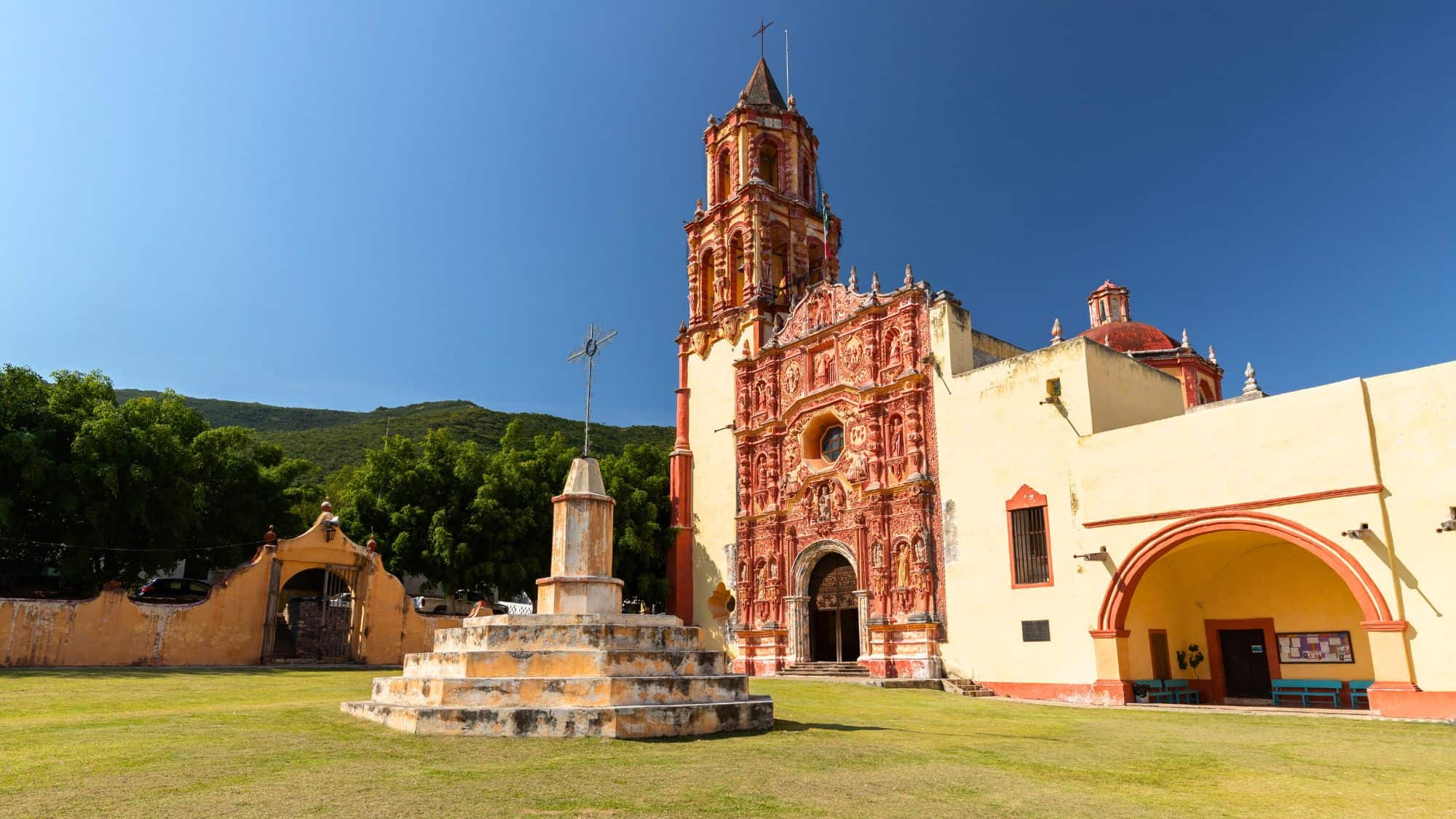 A picturesque colonial-era church in Matamoros, Mexico, with intricate red stone detailing and a bell tower. The surrounding green trees and bright blue sky contrast beautifully with the warm tones of the historic architecture.