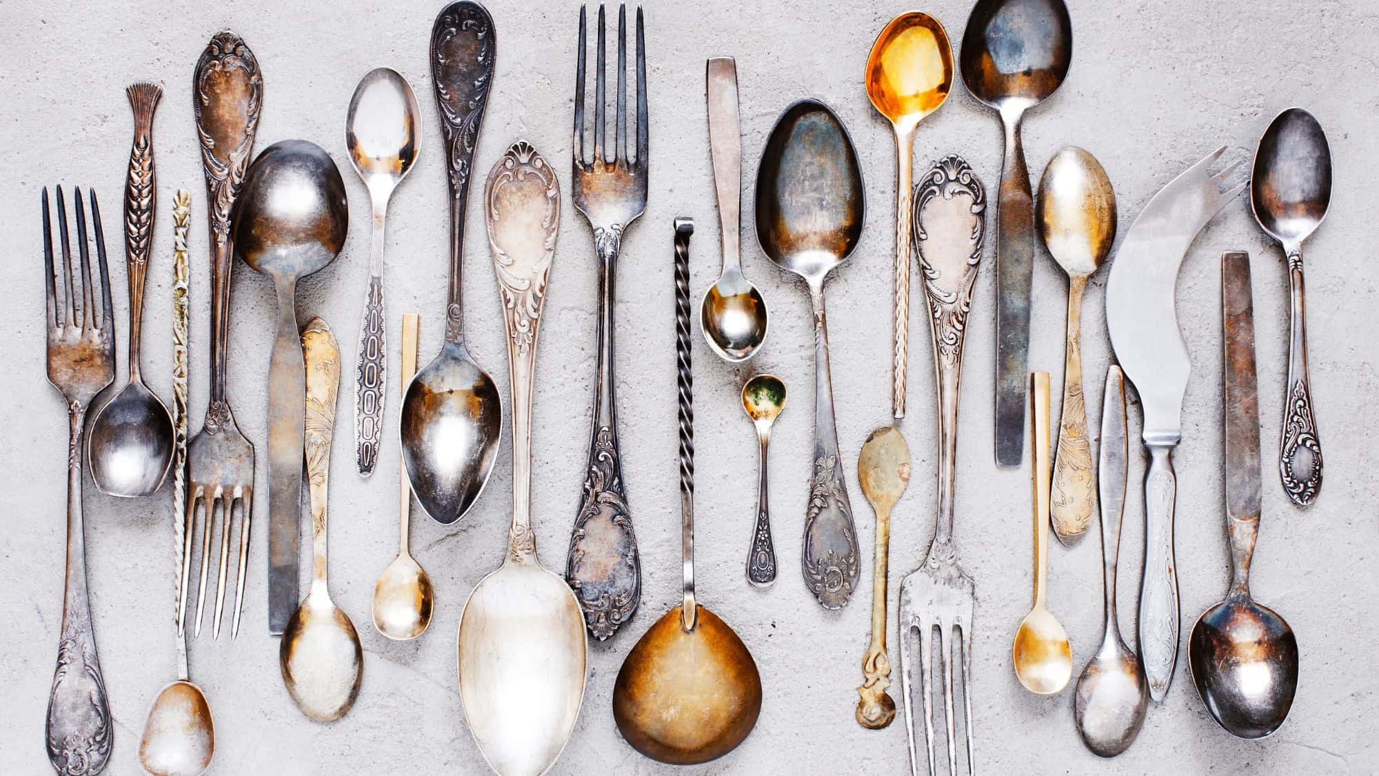 An assortment of antique metal cutlery, including spoons, forks, and knives, displayed on a textured surface. The tarnished silver and gold tones create a rustic, elegant look.