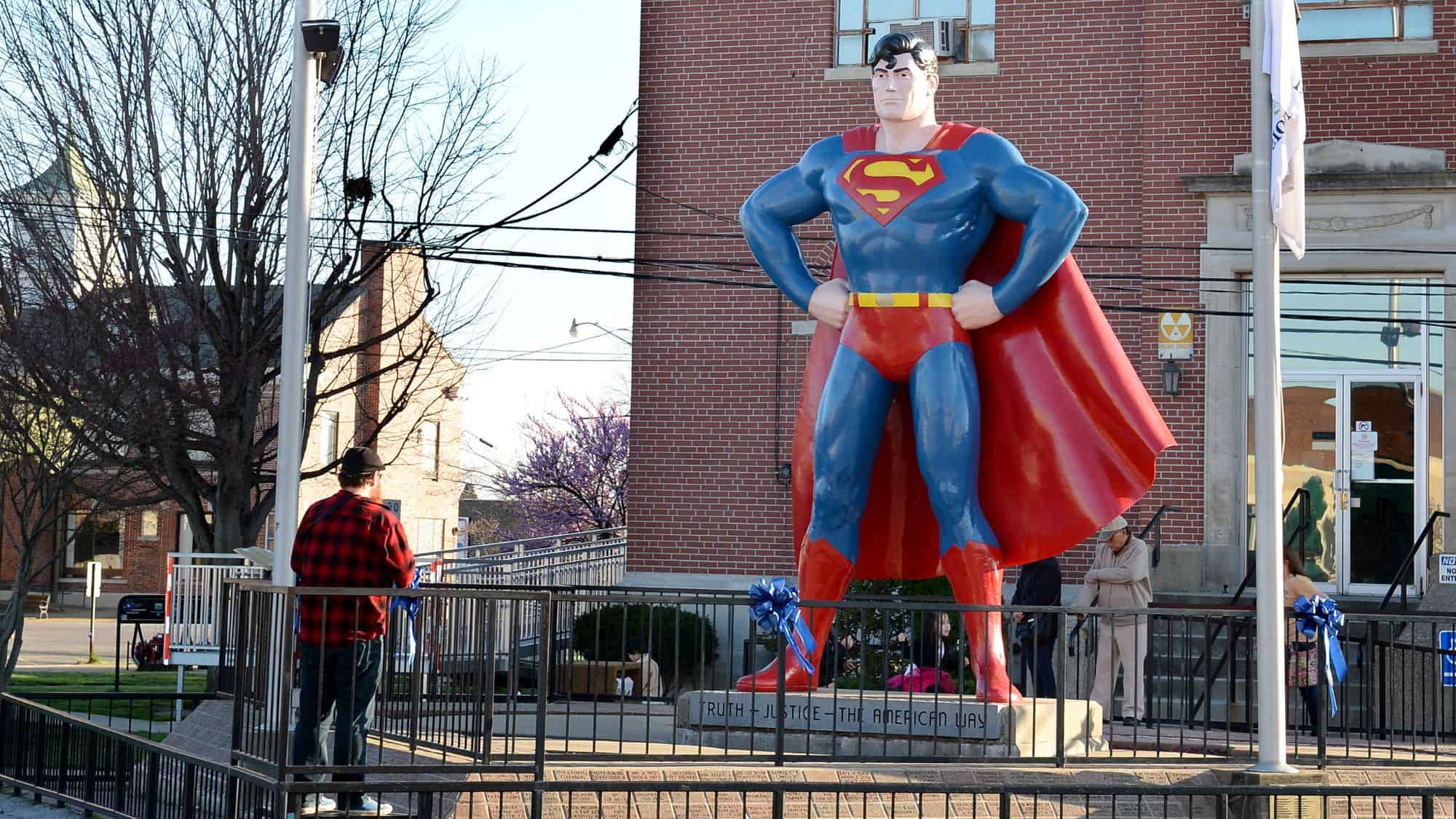 A large statue of Superman standing heroically in front of a red brick building, with his iconic blue suit and red cape. A few visitors stand nearby, taking photos at this famous tribute to the superhero in Metropolis, Illinois.