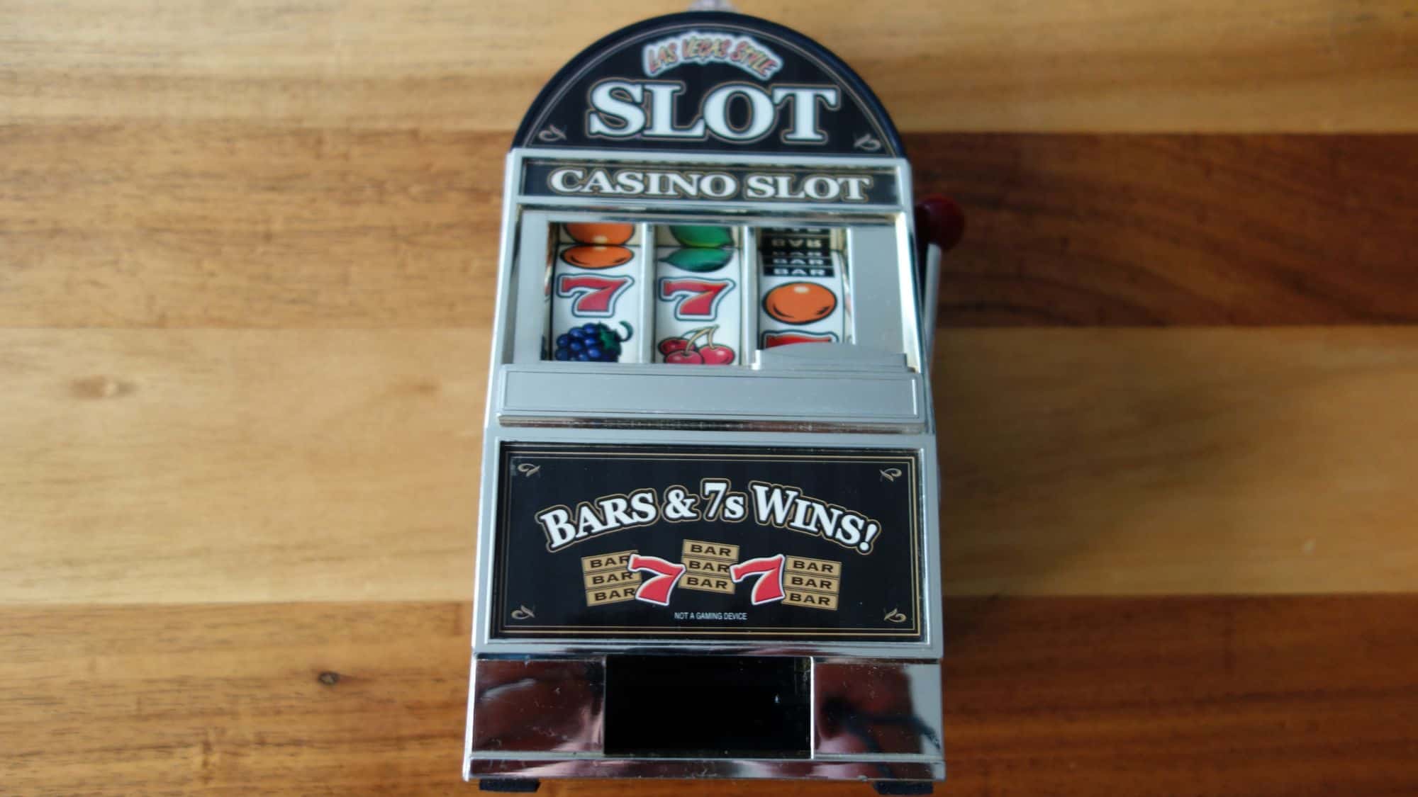A miniature Las Vegas-style slot machine sitting on a wooden surface. The reels display winning symbols like sevens and fruits, with a label reading "Bars & 7s Wins!"
