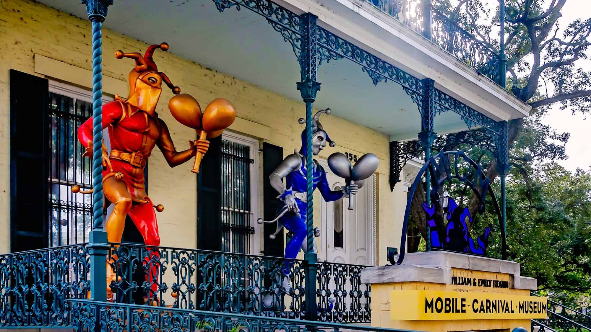 A historic building in Mobile, Alabama, featuring statues of jesters in bright red and blue, holding maracas. The sign reads "Mobile Carnival Museum," celebrating the city’s rich Mardi Gras tradition.