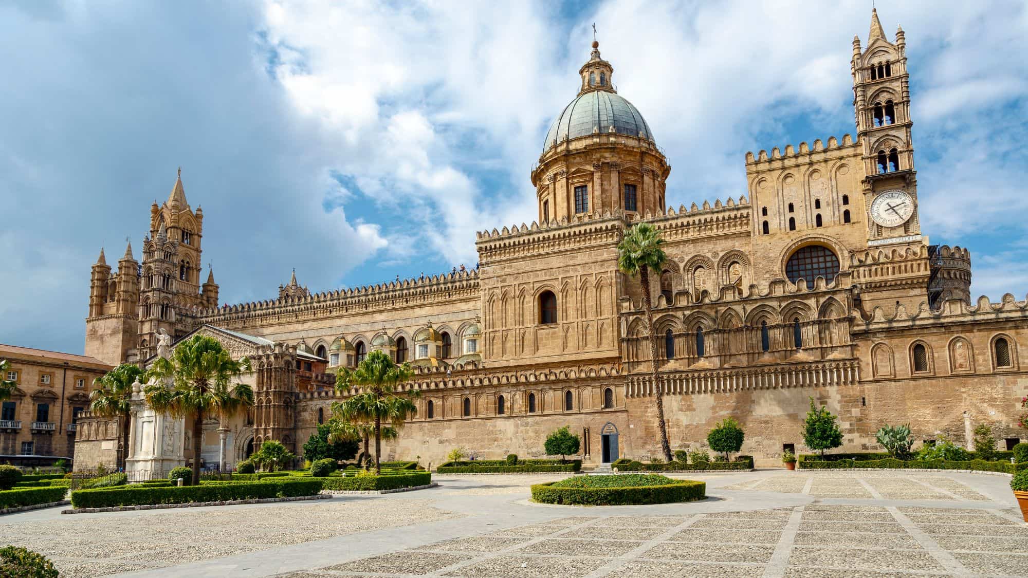 The grand Monreale Cathedral near Palermo, Sicily, showcasing its intricate Norman-Byzantine architecture. The cathedral's dome and towers rise above a lush courtyard, framed by a partly cloudy sky.