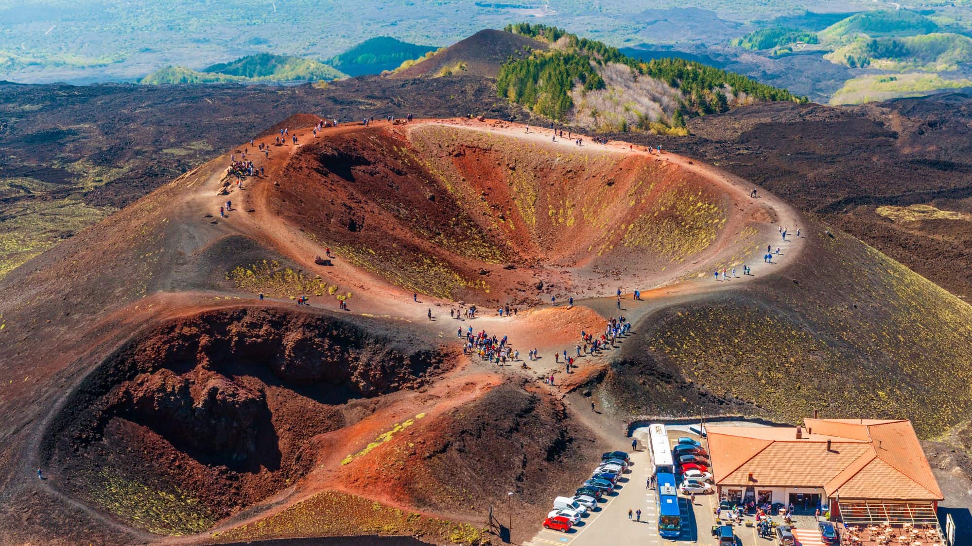 The colorful volcanic craters at the top of Mount Etna, with reddish-brown slopes and patches of green vegetation. Visitors walk along the rim, and a parking area with buses and cars is visible below.