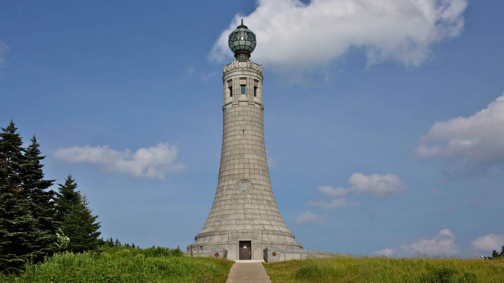 A towering stone monument with a rounded top stands on Mount Greylock under a clear blue sky. The wide grassy field and sparse trees surrounding the monument add to its grandeur.