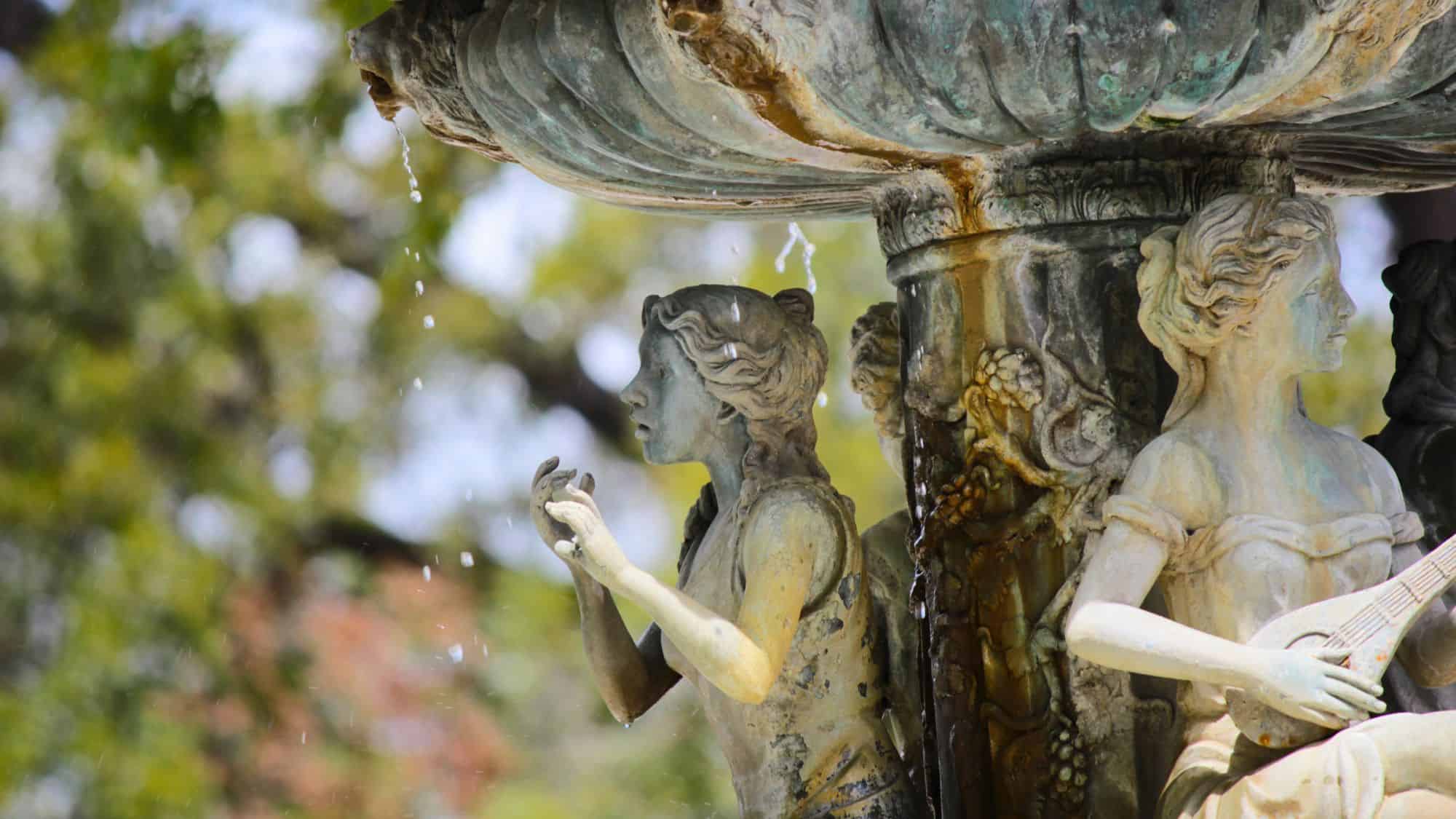 A close-up of an ornate stone fountain in Nuevo Laredo, featuring sculpted figures of women with water droplets falling from the edges. The weathered stone and lush greenery in the background add to the timeless elegance of the scene.