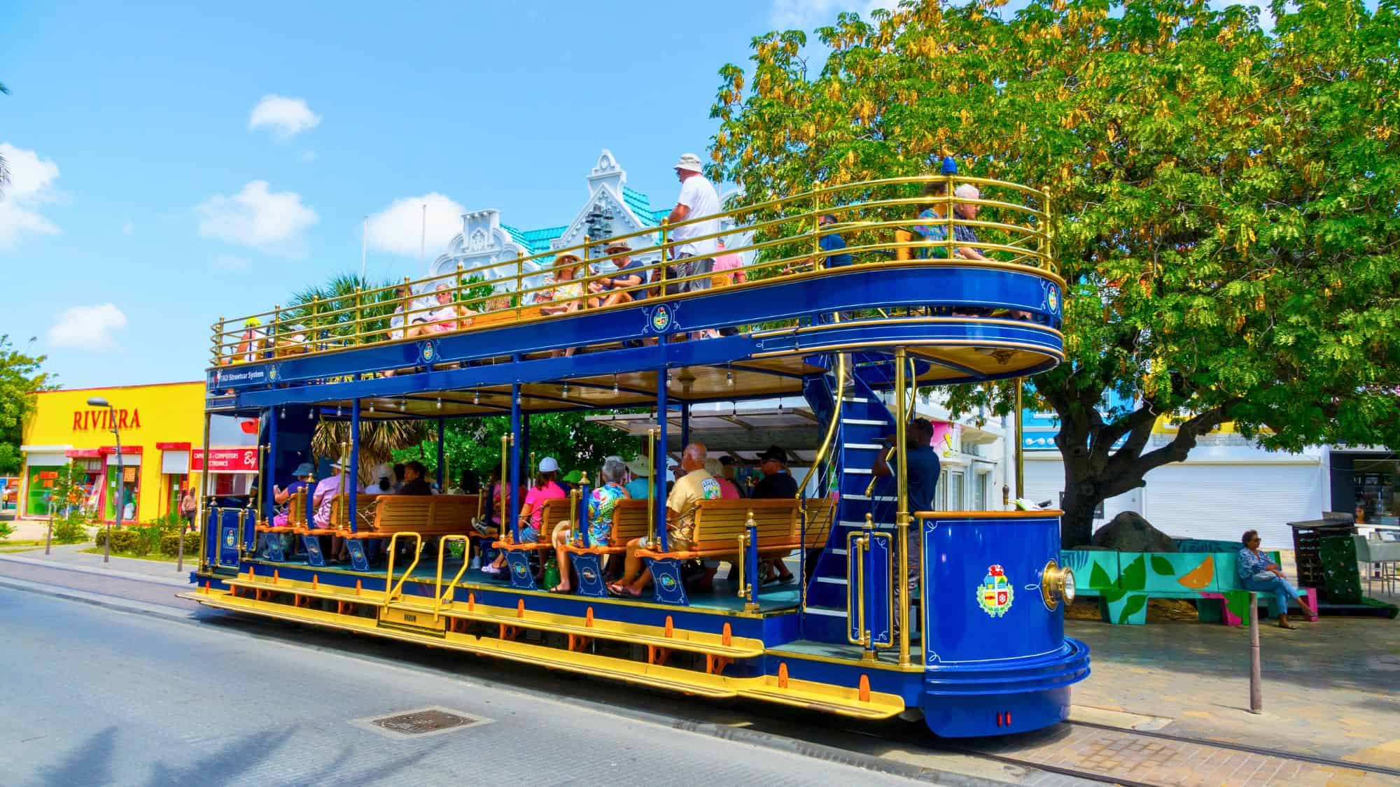 A vibrant double-decker blue and yellow streetcar filled with tourists moves along a colorful street, with shops and tropical trees lining the background under a clear blue sky.