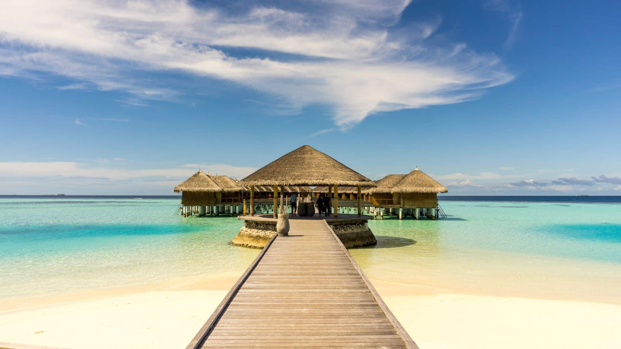 A long wooden boardwalk leads to a cluster of thatched-roof villas perched over clear blue water. The sky is bright and scattered with clouds, enhancing the idyllic island vibe.